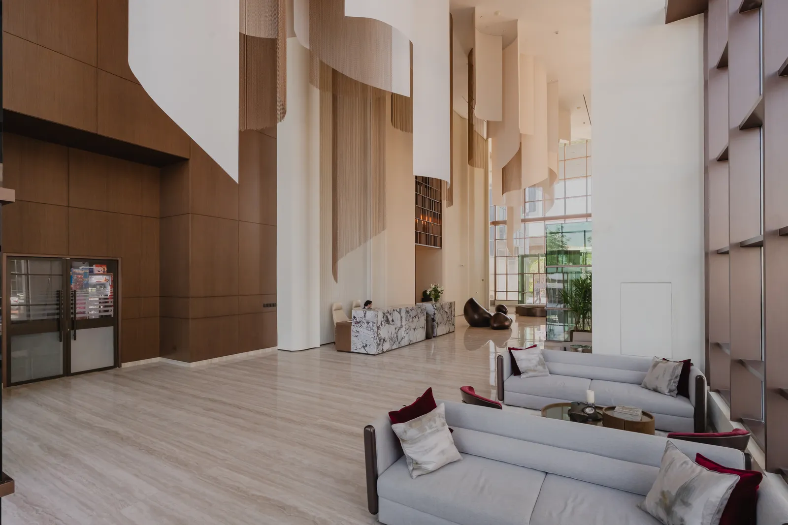 Lobby seating: two grey sofas with burgundy and patterned cushions around round tables, a timber-panelled wall, the hanging fabric sheets, the reception counters and the sculpture beyond