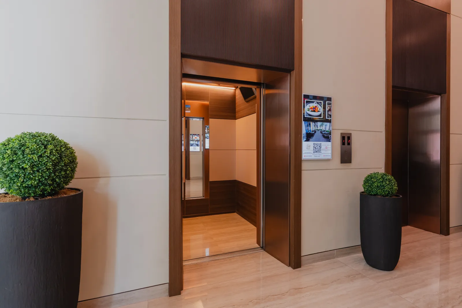 An open lift car with timber panelling, a notice panel beside the call buttons and a second lift door to the right