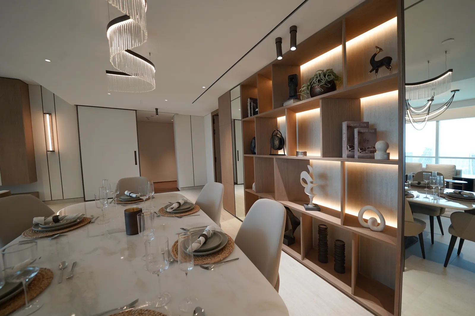 The dining table beside a backlit walnut shelving unit of ornaments, a mirrored wall and the hallway doors beyond