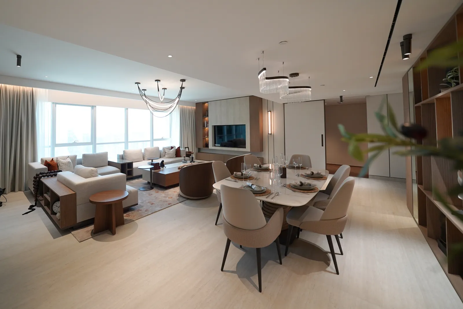 The living room and dining area: cream sofas, a round walnut side table, the dining table under two crystal pendants, the shelving unit and hallway doors