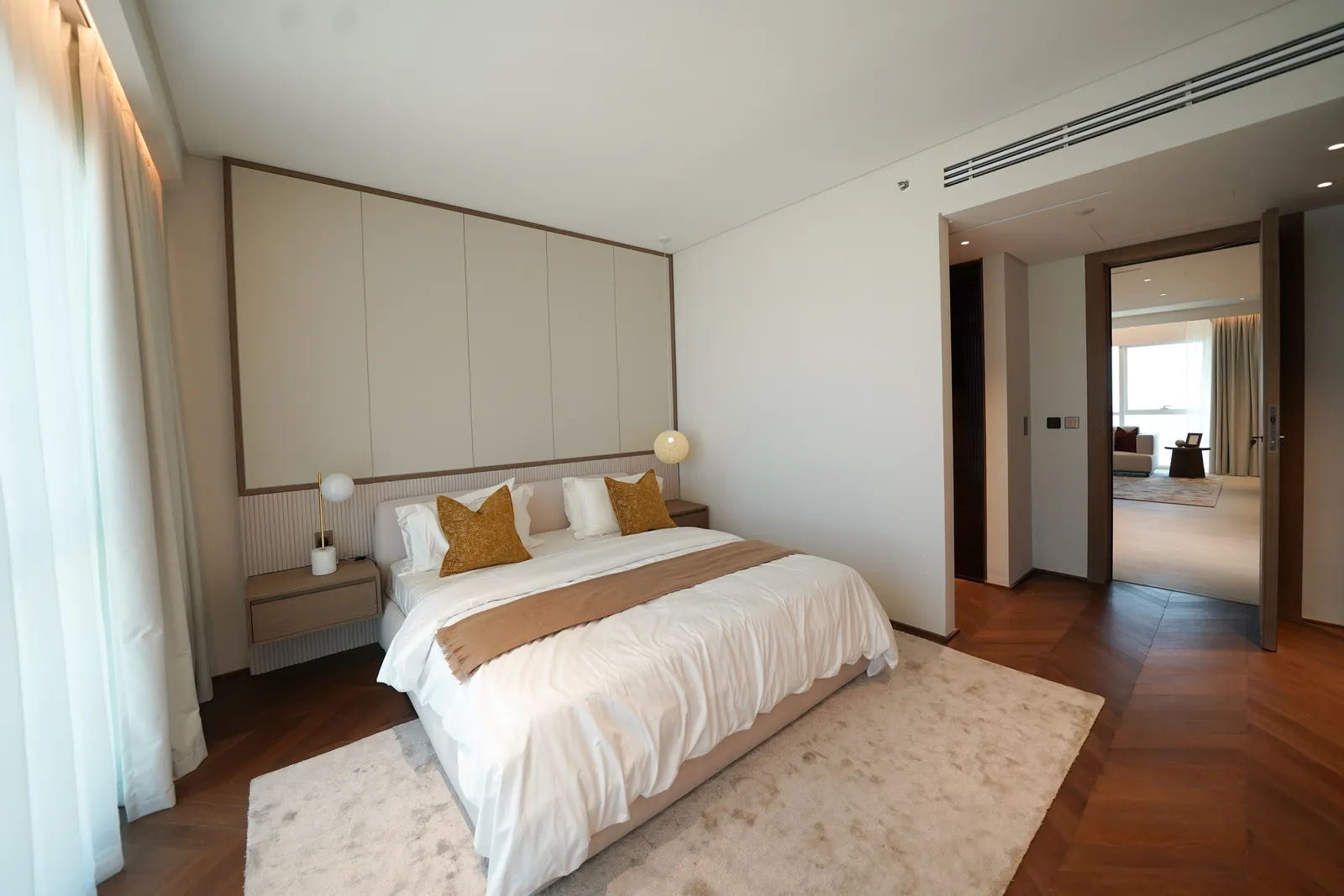A bedroom: a double bed with white linen and mustard cushions against a panelled headboard wall, pendant lights, a herringbone timber floor and a door open to the living room