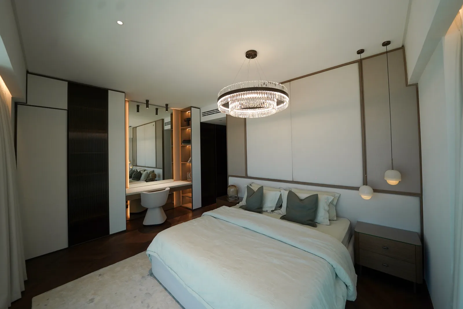 A bedroom with a crystal ring chandelier, a bed with sage cushions, a dressing table with a lit mirror and a white stool, dark wardrobes and a herringbone floor