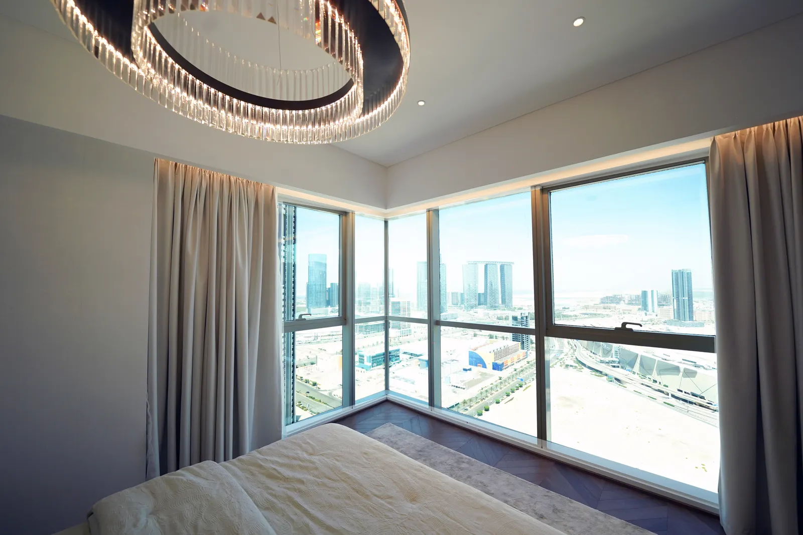 A corner window beside the bed, a crystal ring chandelier above, looking down over Al Reem Island's towers and roads to the water beyond