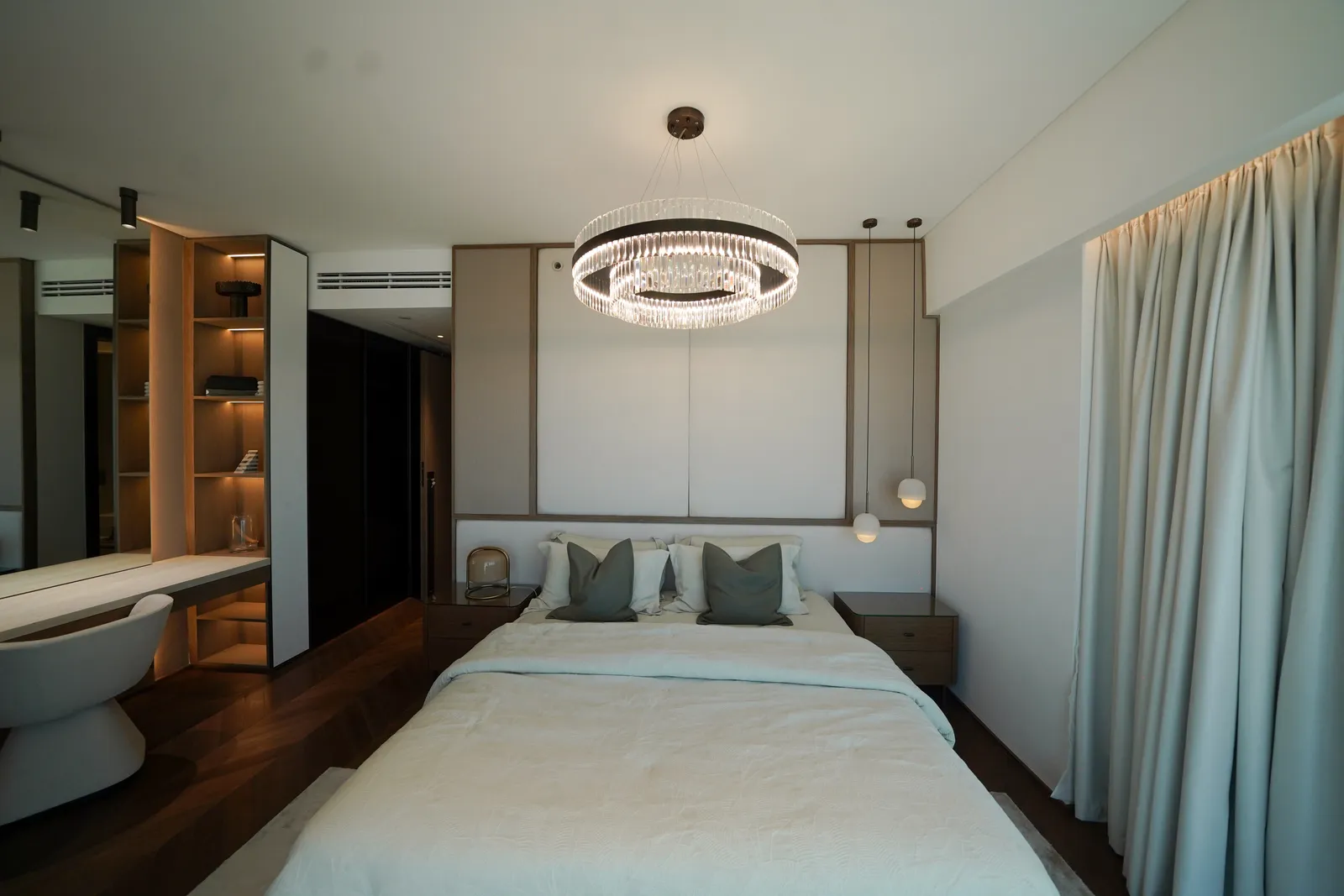 A bedroom: a double bed with grey cushions against a panelled headboard, a crystal ring chandelier, pendant lights and a dressing table with a white stool at left
