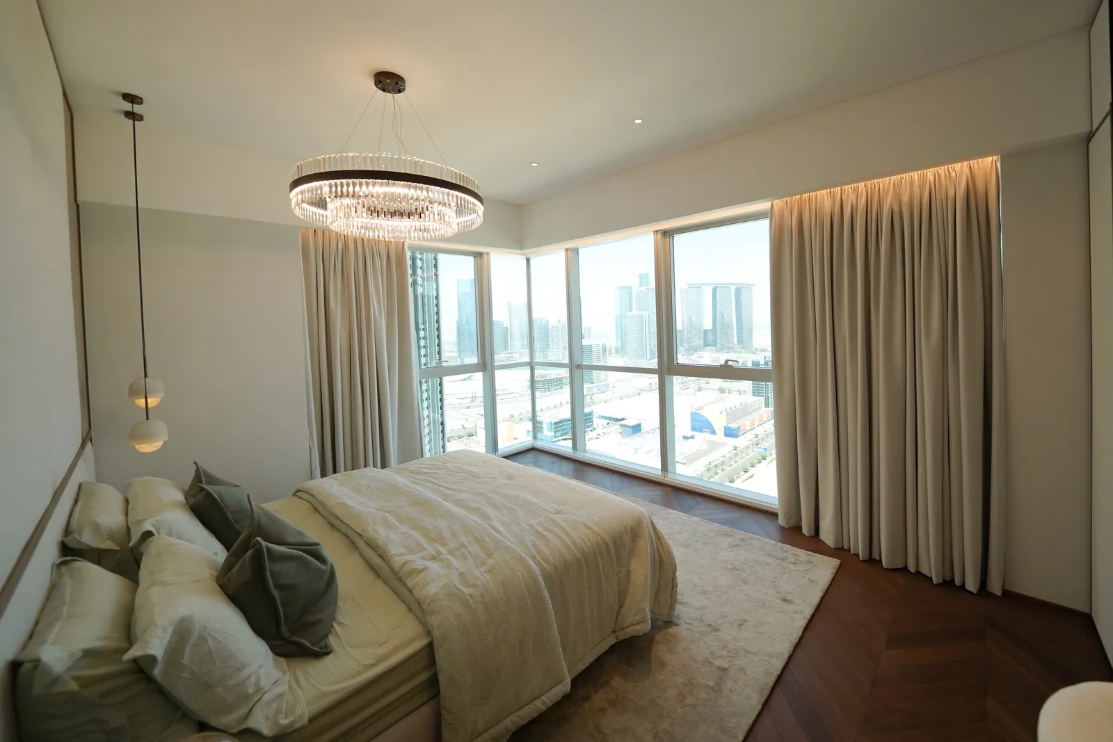 A bedroom with a crystal ring chandelier, a glass pendant light, curtains drawn back from the corner window and a herringbone floor