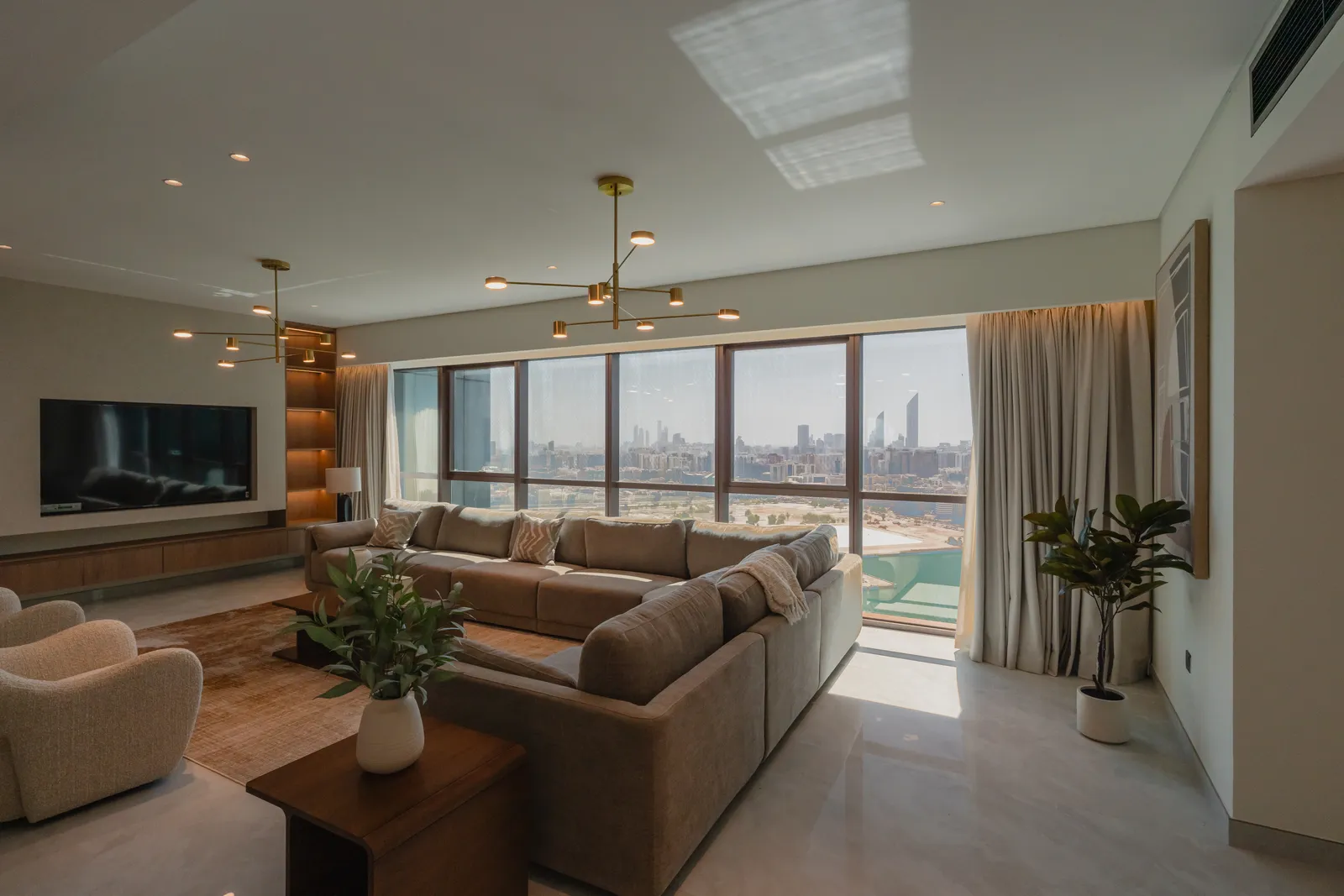 The furnished living room by day: the tan sectional sofa, a television wall with lit shelving, brass branched lights and the skyline across the water through the windows