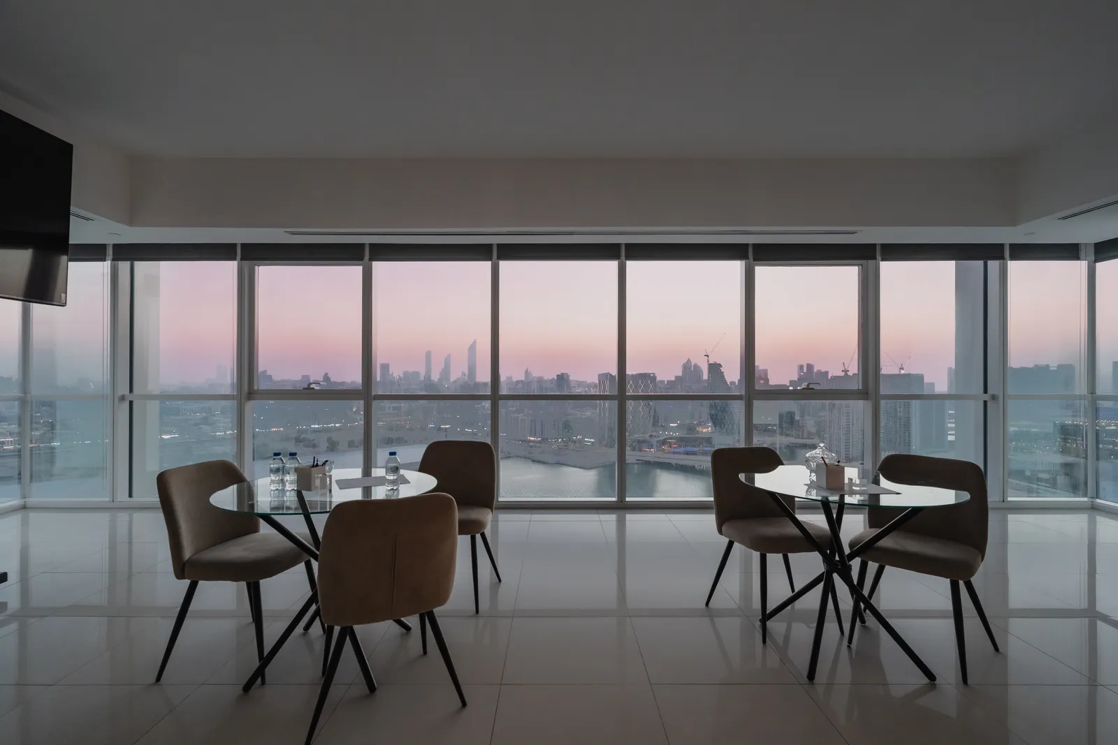 An apartment at dusk with two small tables and chairs by the glazing, the pink sky and the city skyline across the water beyond