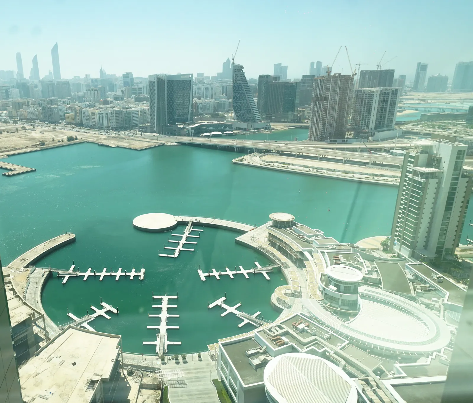 The view down to the marina by day: pontoons with moored boats, a round-roofed waterfront building, towers under construction across the channel and the skyline beyond