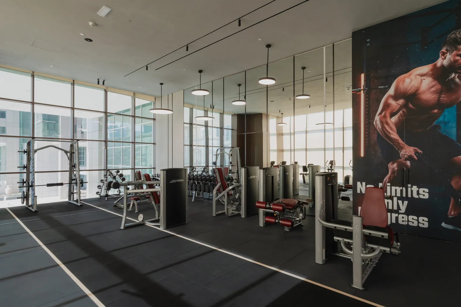 The gym: a Smith machine, benches and strength machines on a dark floor by the glazed wall, under a wall-sized poster reading No Limits Only Progress