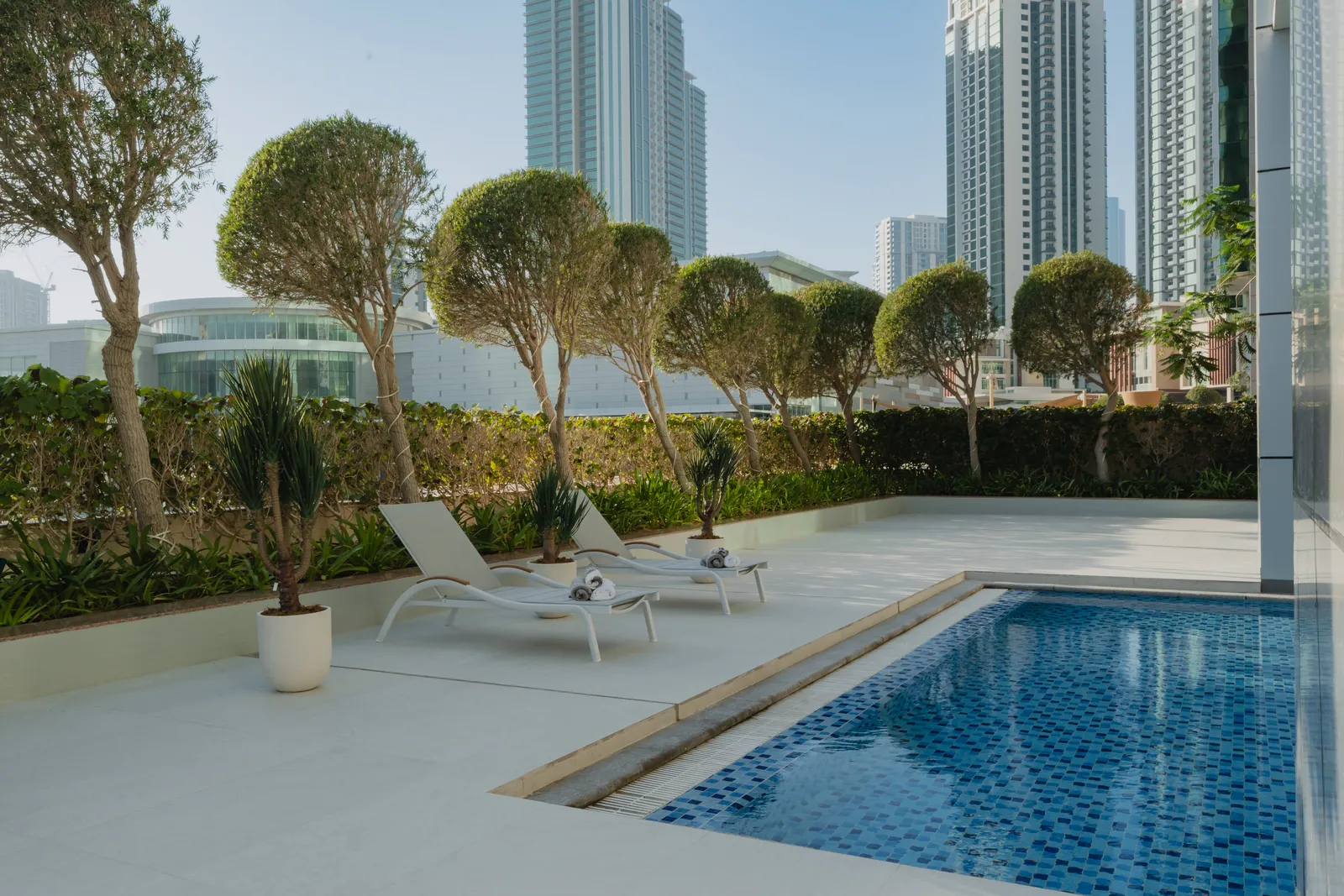 The children's pool: a small mosaic-tiled pool beside two loungers with towels, potted plants, a row of clipped trees above the hedge and Al Reem Island's towers beyond