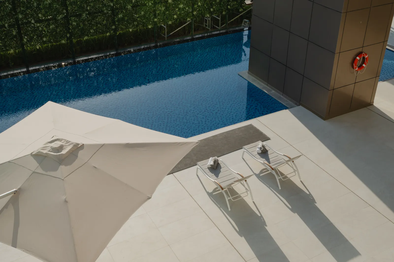 The pool from above: a cream parasol, two loungers with towels on a mat, the bronze column with a lifebuoy and the hedge screen