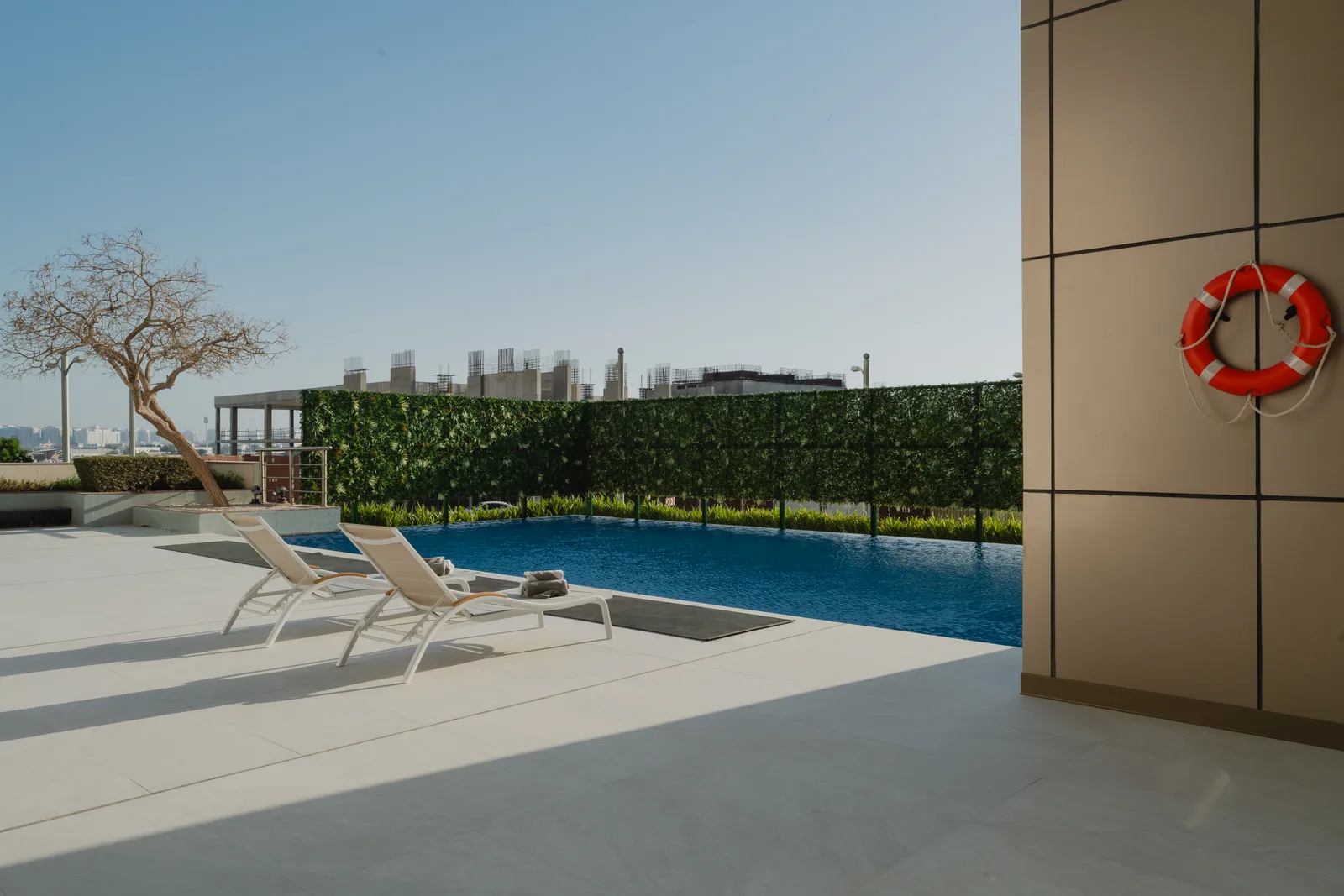 Two loungers with towels at the poolside, the hedge screen, a bare tree and buildings under construction beyond, a lifebuoy on the bronze wall