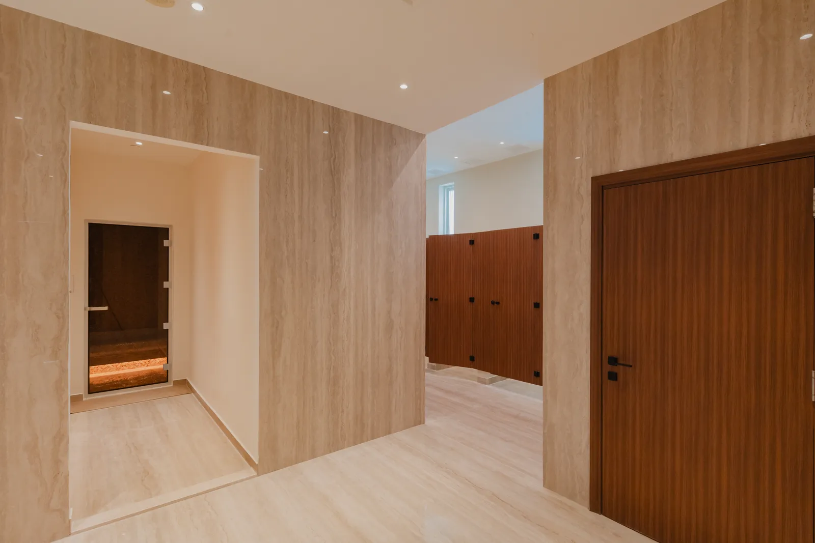 The changing room: travertine-clad walls, the glass door of the sauna at left, timber lockers and cubicle doors beyond
