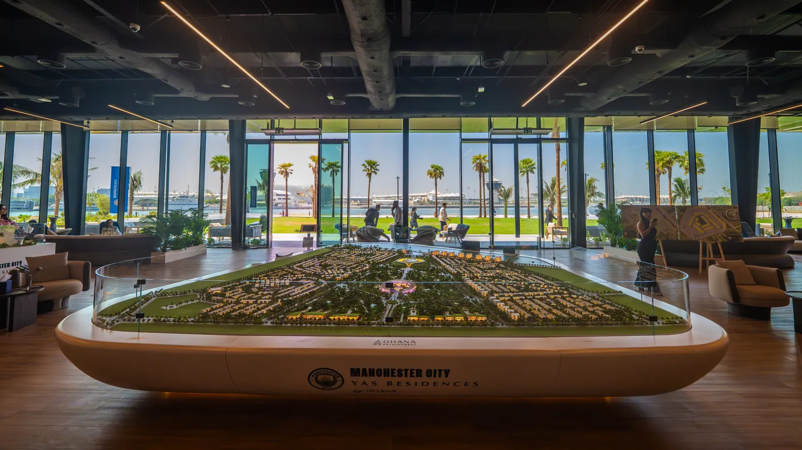 The community’s scale model on a curved white plinth in the sales centre, the canal and palms through the glass wall behind, lounge seating either side