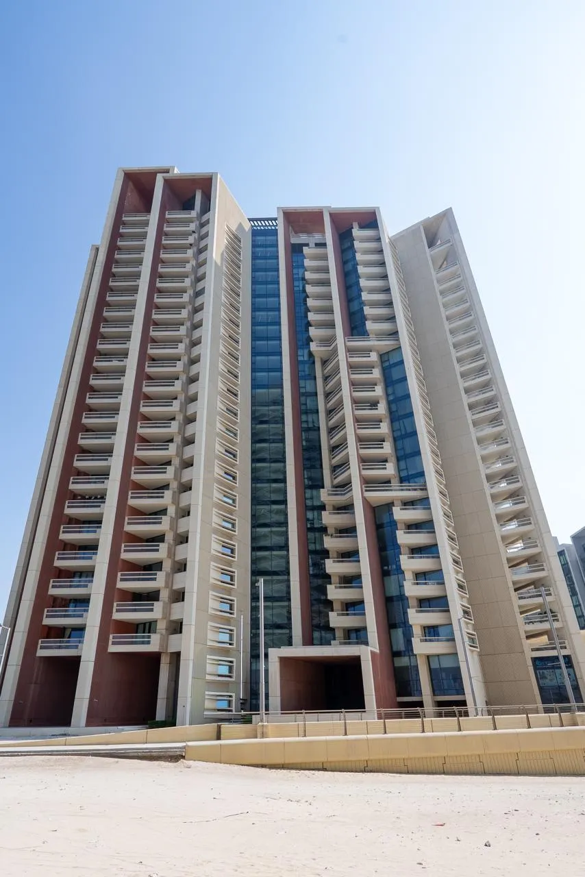 Marafid Tower in portrait from the sand beside it: the full height of the tower, balconies stacked between terracotta fins, the podium wall at the base