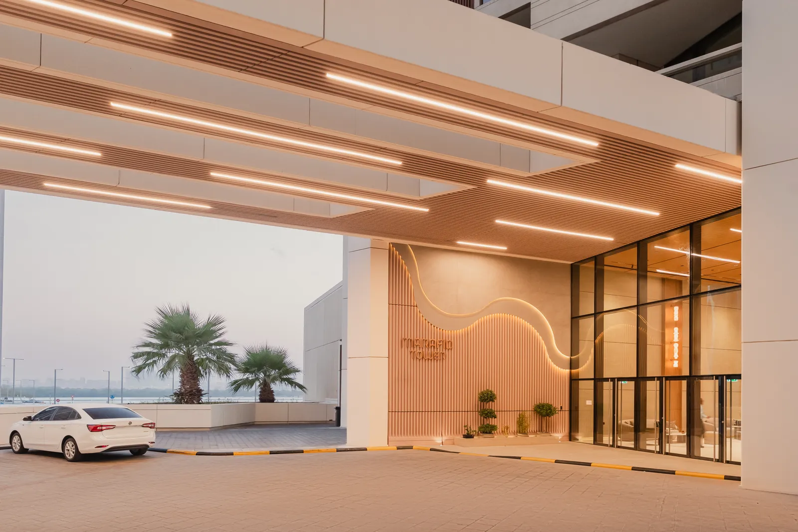 The entrance at dusk: a lit canopy over the drop-off, a fluted wall with a wave of light and the MARAFID TOWER sign beside the glass doors, palms and a parked car outside