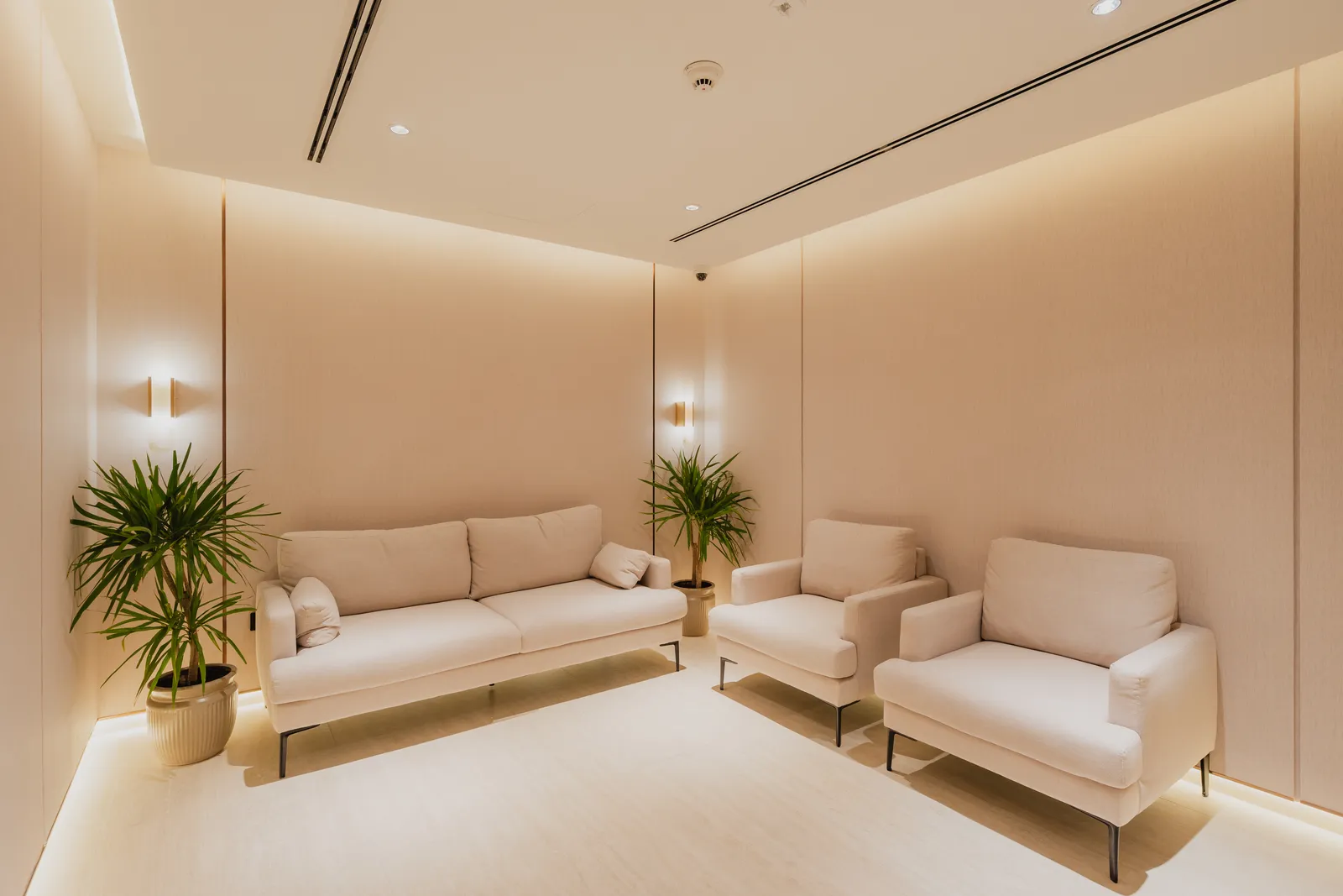 A residents' lounge: a cream sofa and two armchairs on brass-framed bases, two potted palms and a pair of wall lights on a plain cream wall