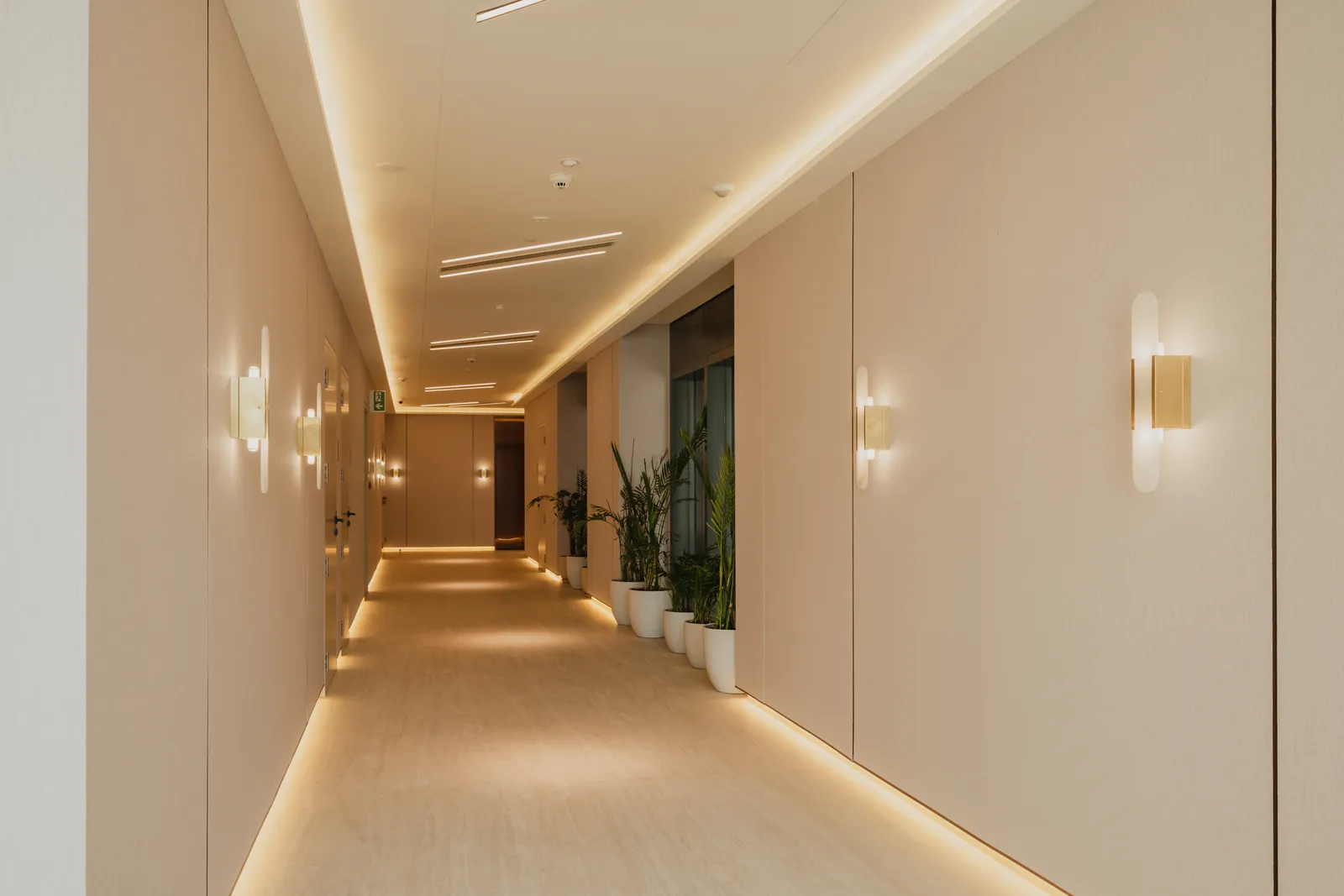 A residential corridor: apartment doors on both sides, brass wall lights, a plant and a long run of floor-level light
