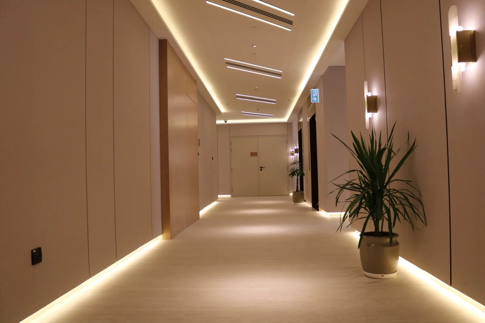 A residential corridor with cove lighting along the ceiling, wall lights and a potted plant